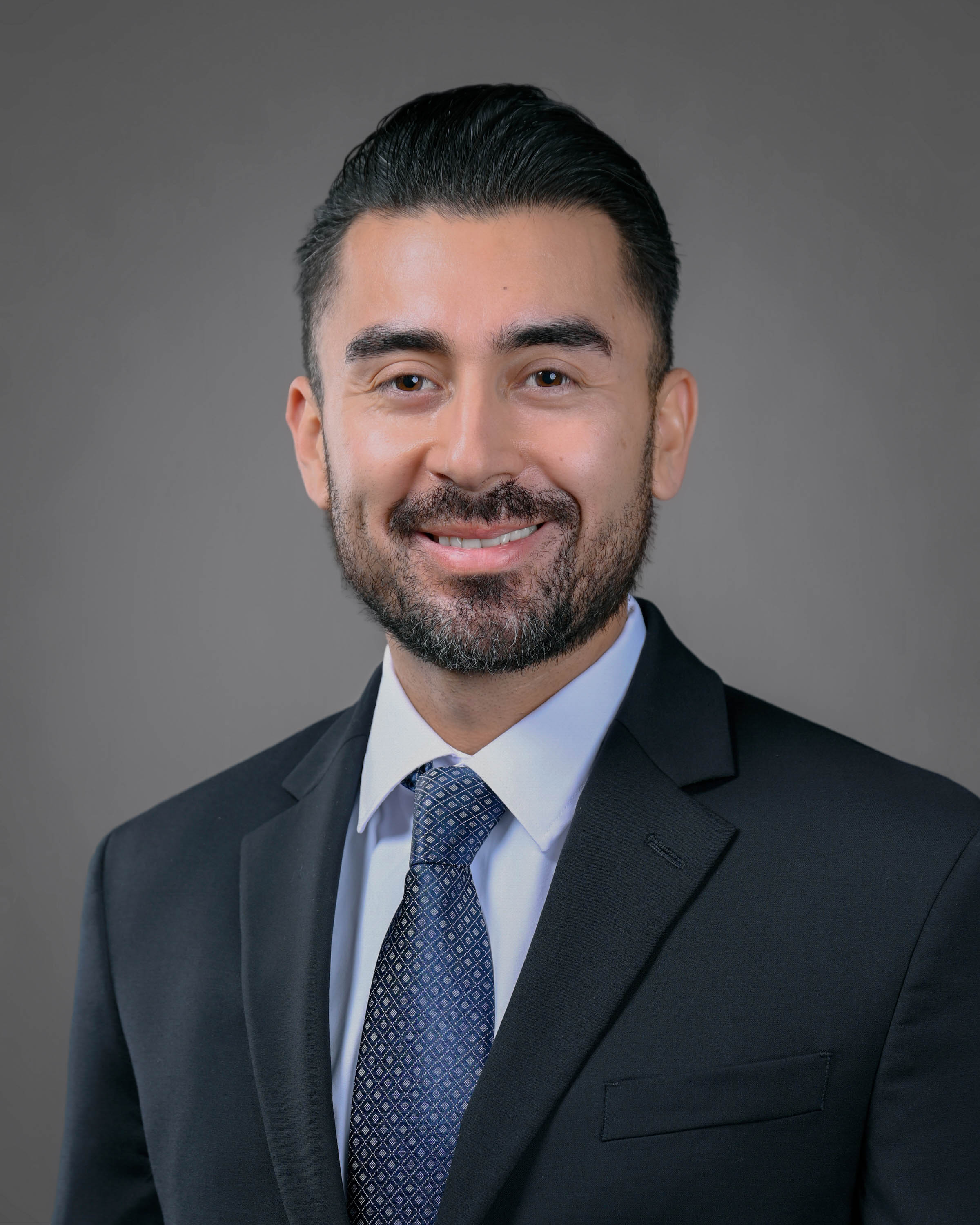 Man with dark hair and a short beard smiling in a dark blue suit with a blue dotted tie